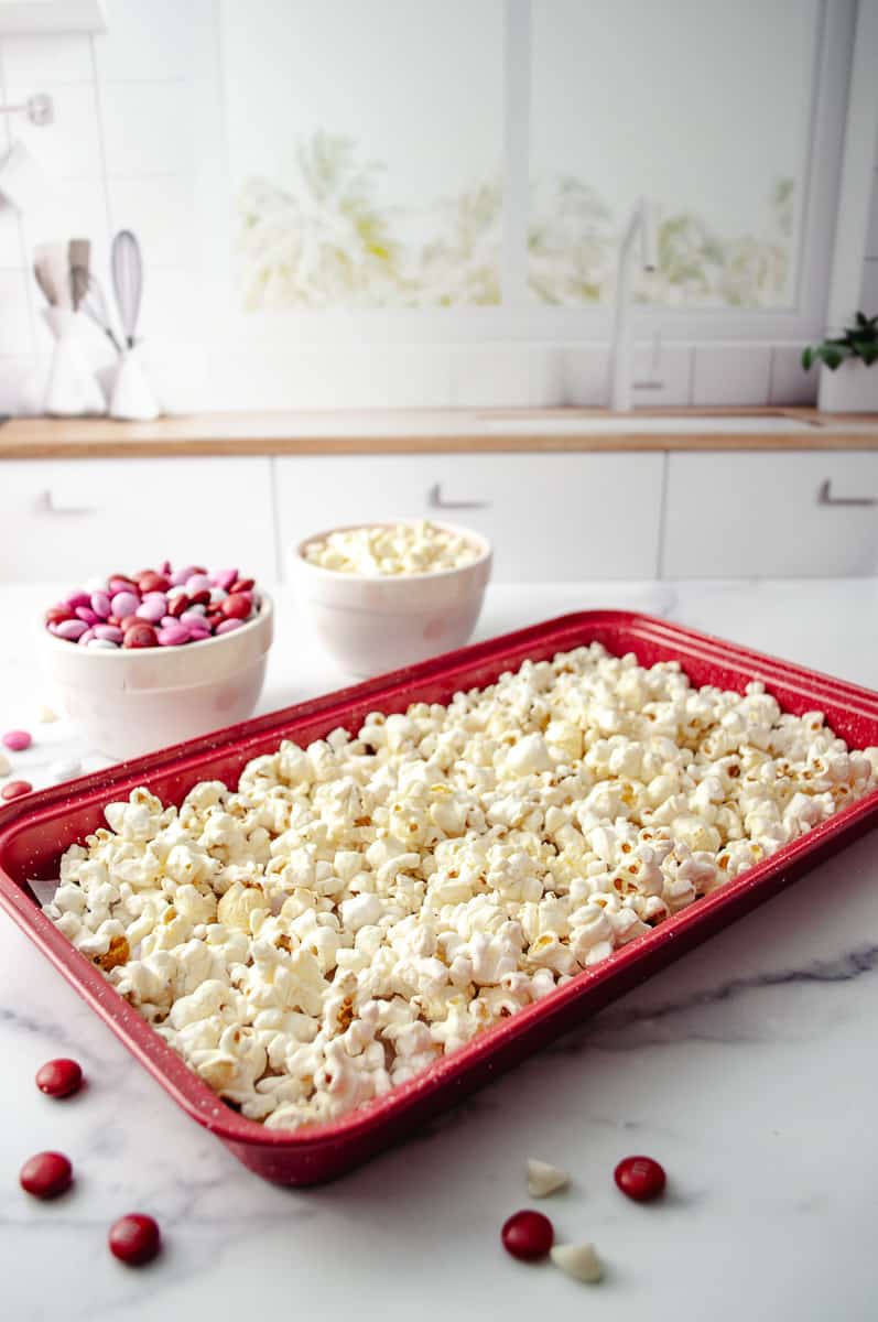 Easy Valentine's White Chocolate M&M Popcorn Treat  popcorn in a red tray with two small bowl in the background with valentines m&ms and white chocolate chips. 