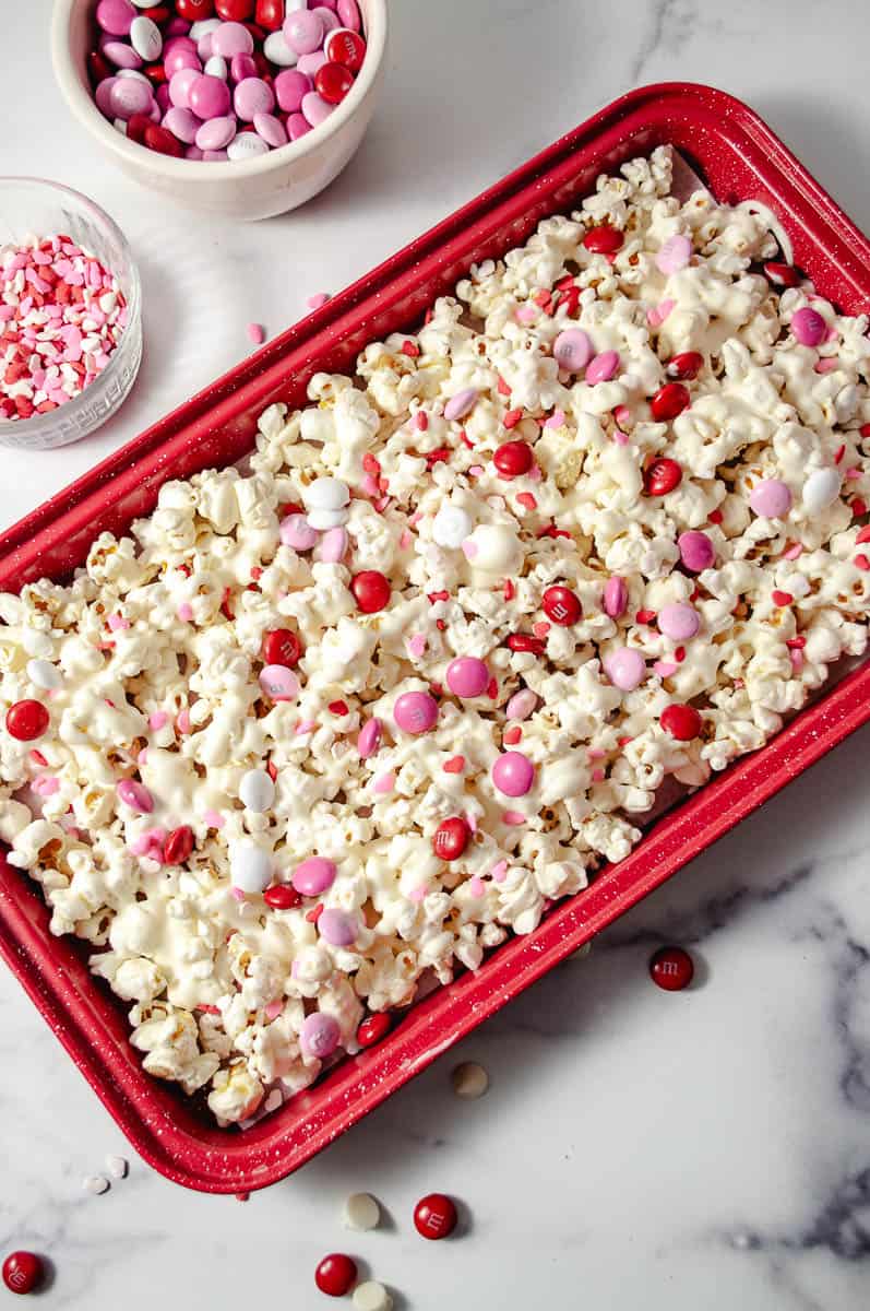 Easy Valentine's White Chocolate M&M Popcorn Treat  the finished popcorn creat in the red try with a bowl of m&ms and a mini clear bowl of red, pink and white heart sprinkles. 