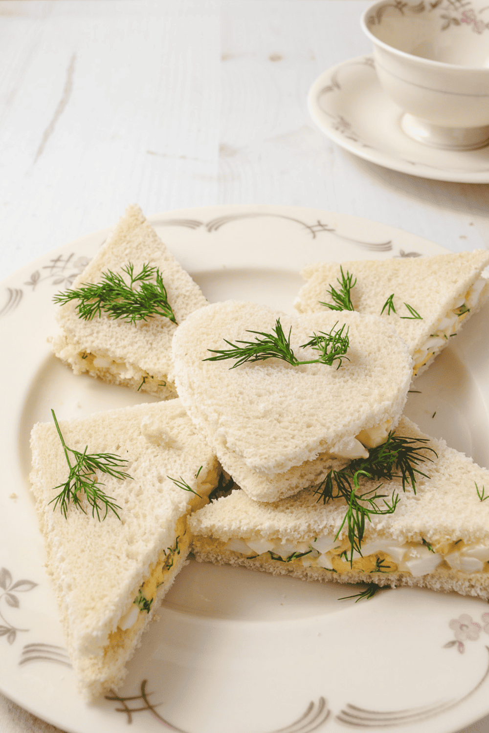 Tea party Sandwiches 3 triangles and one heart with eg salad with dill springs on top on a cream plate with a tea cup and saucer on a white table. 