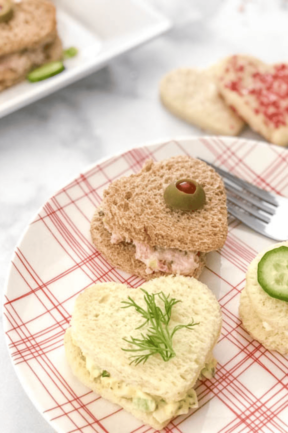 Heart shape tea sandwiches on a white and red plan played with a fork to heart cookies Anna White counter and the corner of another platter with a heart shaped tea sandwich.