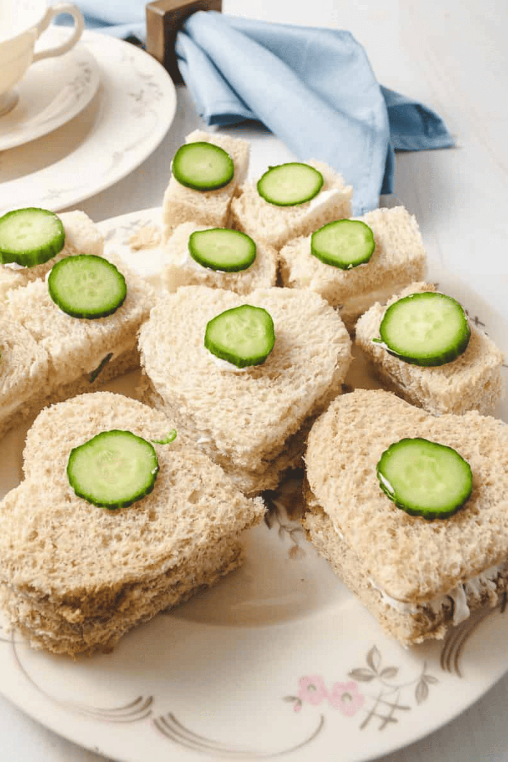 Perfect Party Tea Sandwich 3 Easy Filling Recipes 3 heart shaped with cream cheese filling and small slices of cucumber on top and 8 square one on a white table with a blue napkin.