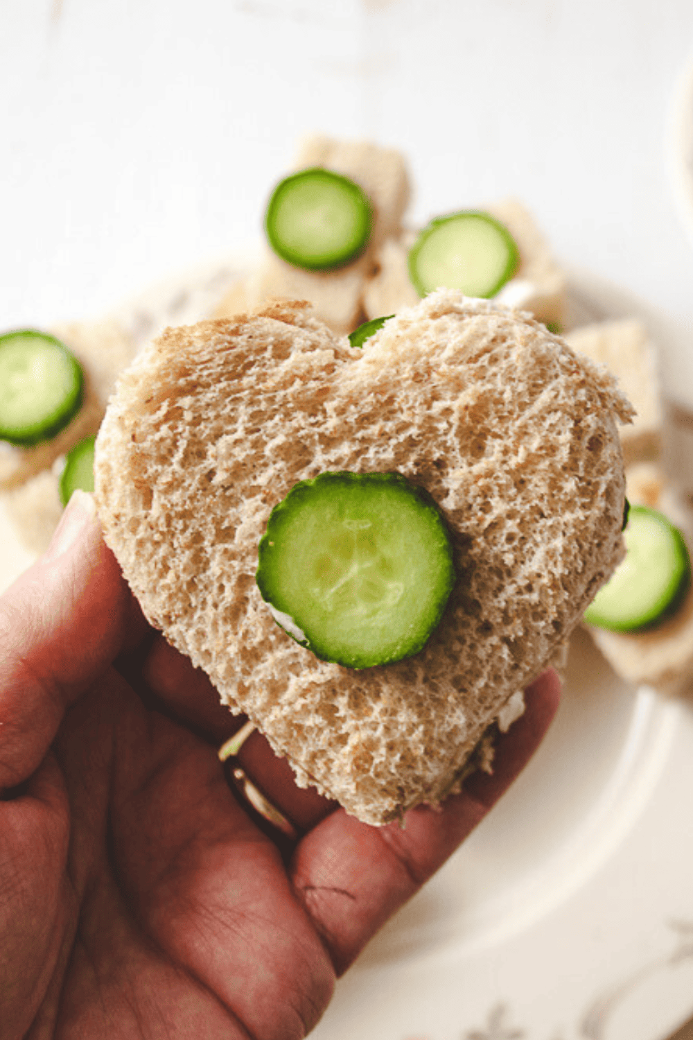 Close up of the heart shaped tea sandwich in a hand with a plate with other sandwiches in the blurred background