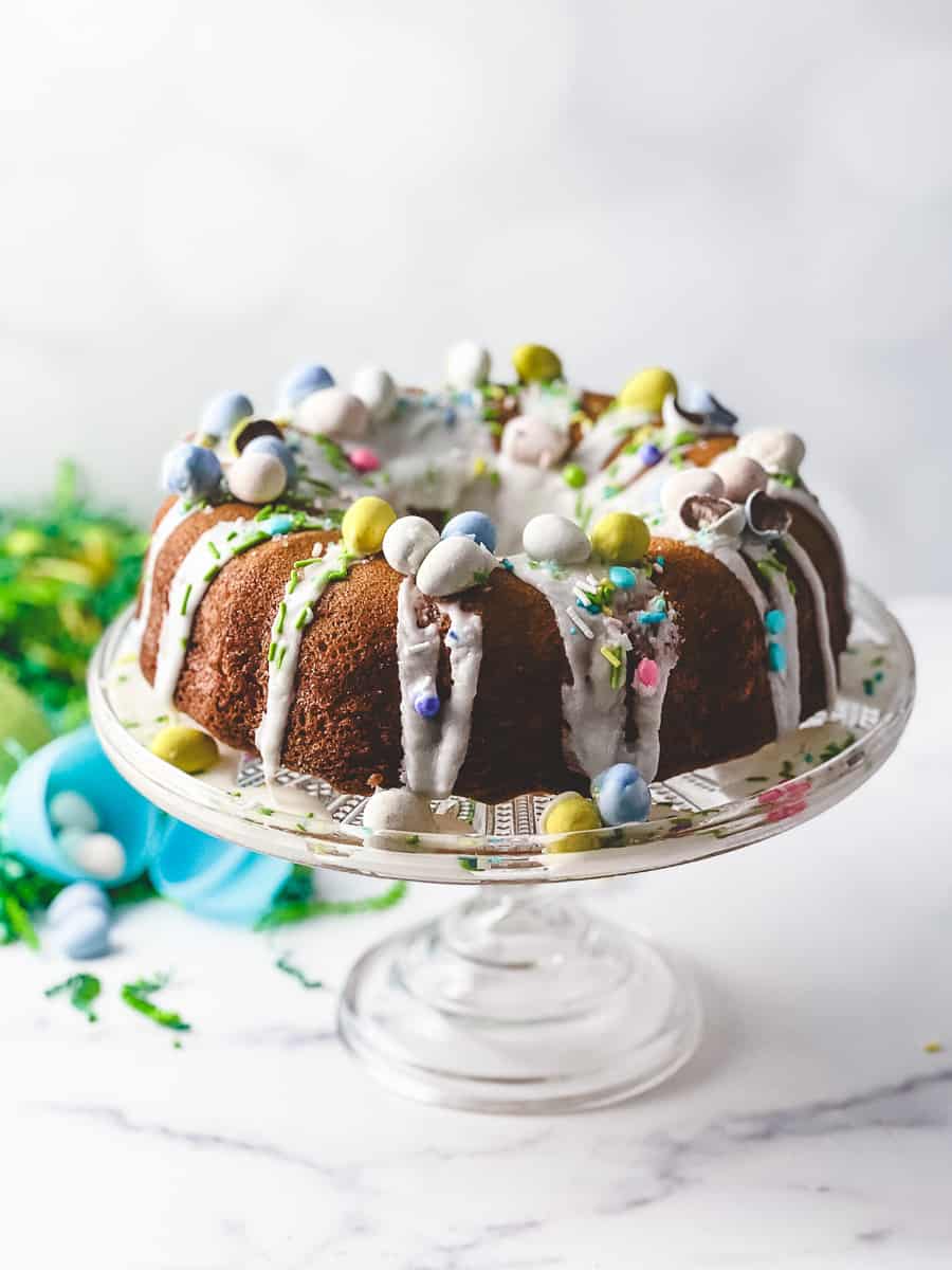 Easy Easter Lemon Bundt Cake: Box Cake Mix Recipe on a short clear glass cake stand with green easter grass and a blue plastic easter egg with cadbury mini eggs on the counter and all over the cake with a white lemon drizzle.