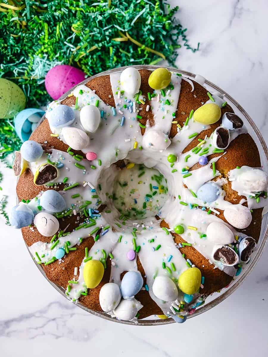 top down view Easy Easter Lemon Bundt Cake: Box Cake Mix Recipe on a short clear glass cake stand with green easter grass and a blue plastic easter egg with cadbury mini eggs on the counter and all over the cake with a white lemon drizzle. 
