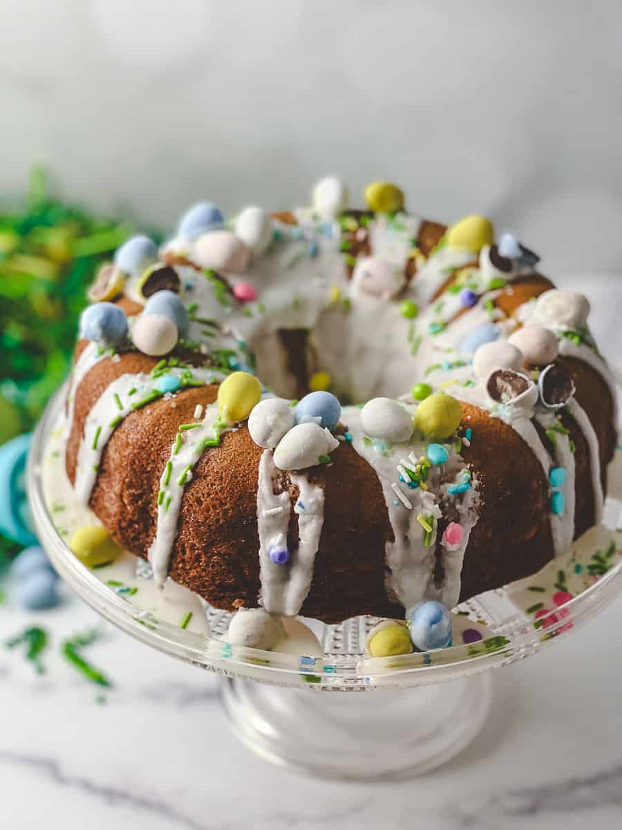close up view of the top of the cake Easy Easter Lemon Bundt Cake: Box Cake Mix Recipe on a short clear glass cake stand with green easter grass and a blue plastic easter egg with cadbury mini eggs on the counter and all over the cake with a white lemon drizzle. 