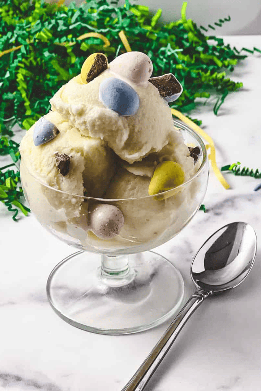 The Best Ninja Creami Vanilla Protein Ice Cream creamy ice cream in a parfait glass with a spoon cadbury mini eggs mixed in and green easter grass in the background.