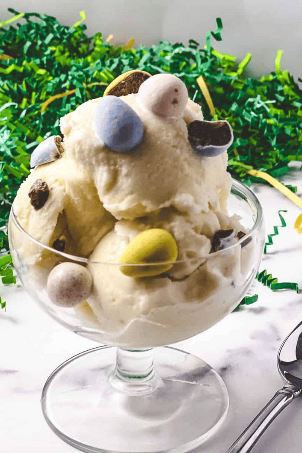 close up of The Best Ninja Creami Vanilla Protein Ice Cream creamy ice cream in a parfait glass with a spoon cadbury mini eggs mixed in and green easter grass in the background.