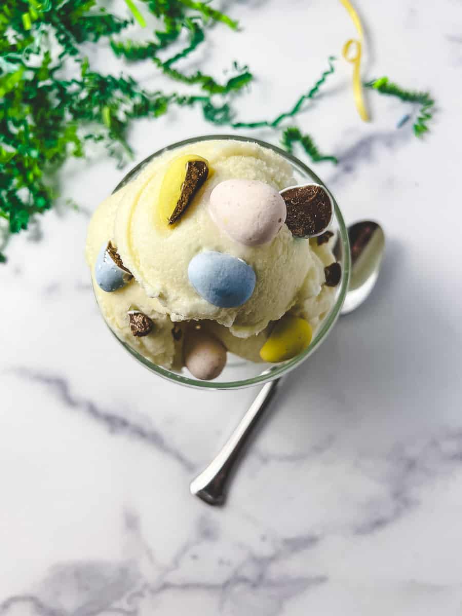 top vie of The Best Ninja Creami Vanilla Protein Ice Cream creamy ice cream in a parfait glass with a spoon cadbury mini eggs mixed in and green easter grass in the background.