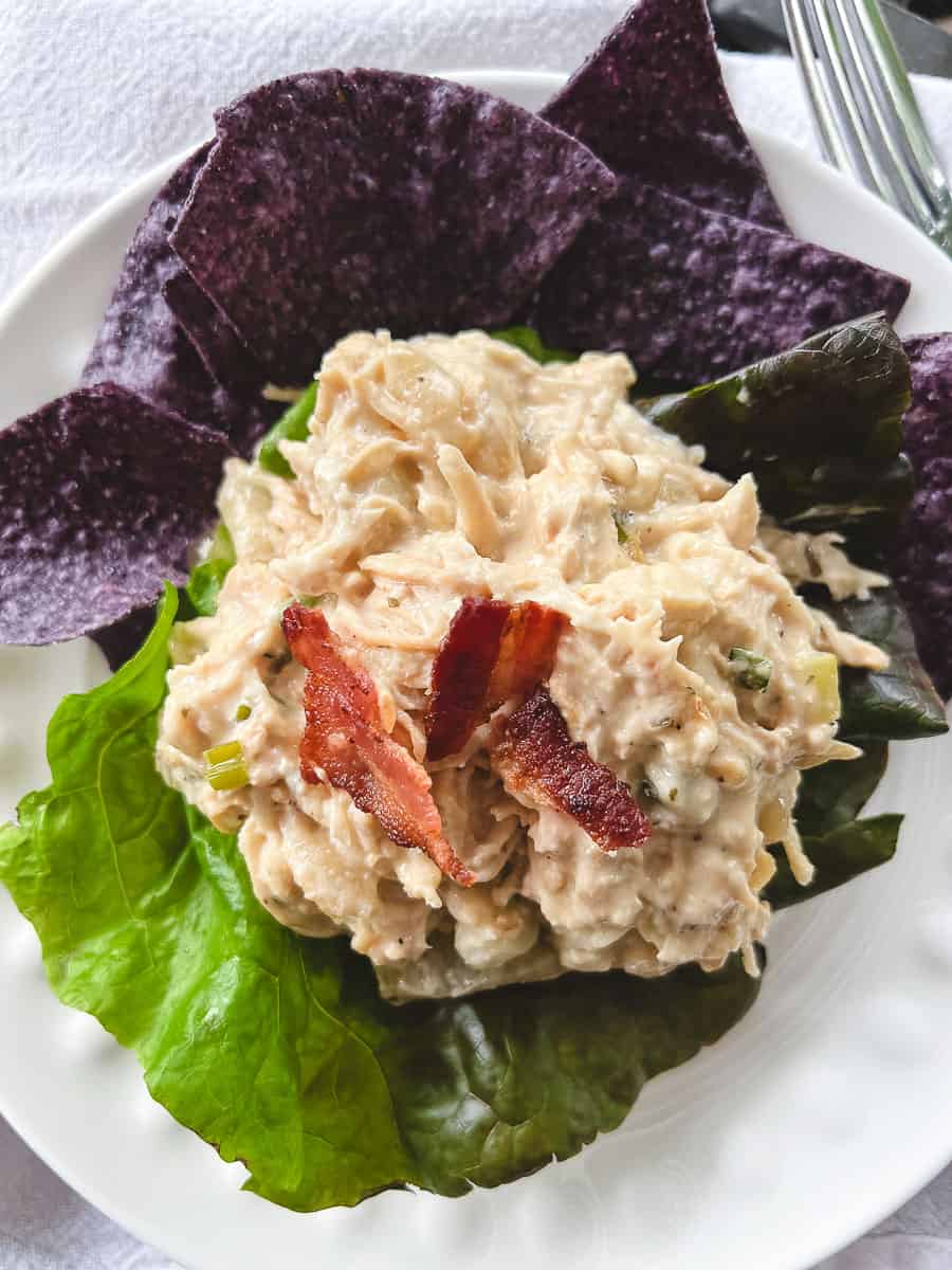 Slow Cooker Cream Cheese Crack Chicken Bacon and Ranch on a small round white plate on a lettuce leaf with blue corn chips on the plate and a fork on the right side. 