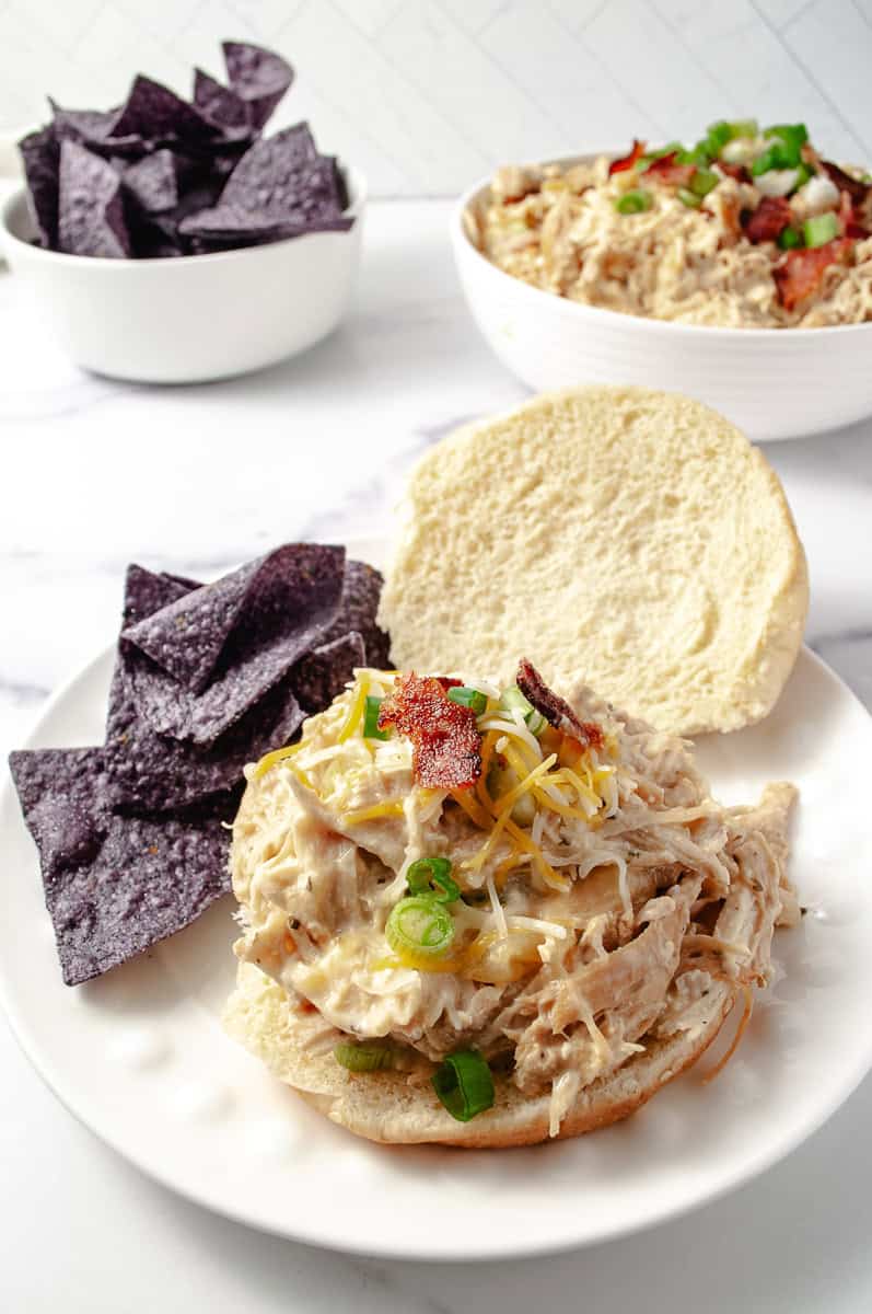 Slow Cooker Cream Cheese Crack Chicken Bacon and Ranch on a bun on a round white plate with blue corn chips on the plate and in the background in white bowls blue corn chips and the chicken in a serving bowl. 