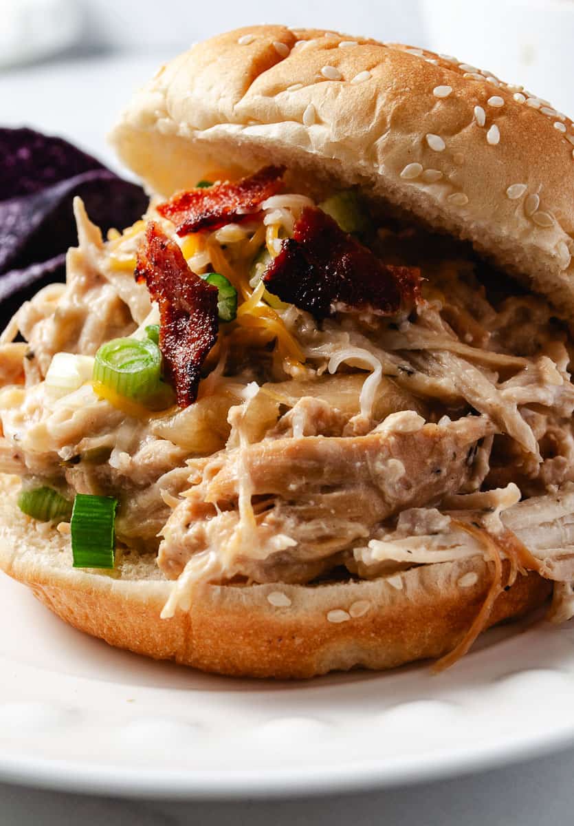 Slow Cooker Cream Cheese Crack Chicken Bacon and Ranch. on a bun with sesame seeds on top and blue corn chips on the plate.