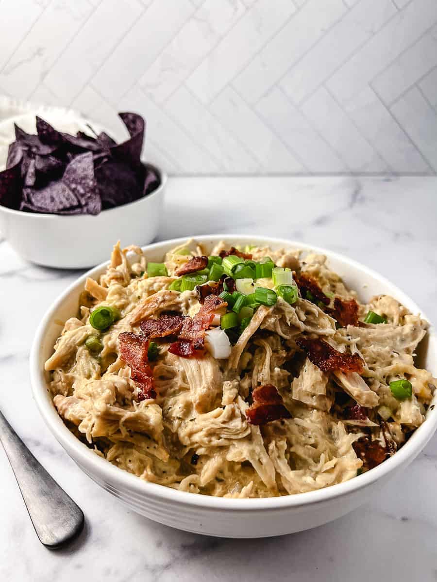 Slow Cooker Cream Cheese Crack Chicken Bacon and Ranch in a round white serving bowl with a small white bowl with blue corn chips and a spoon on the left side.