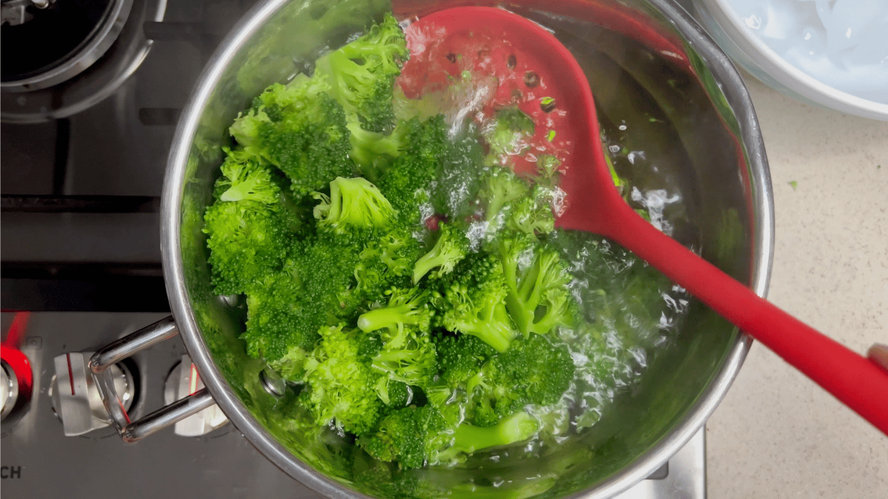 parboiling broccoli in a saucepan on the stove with a red slotted spoon removing the broccoli.
