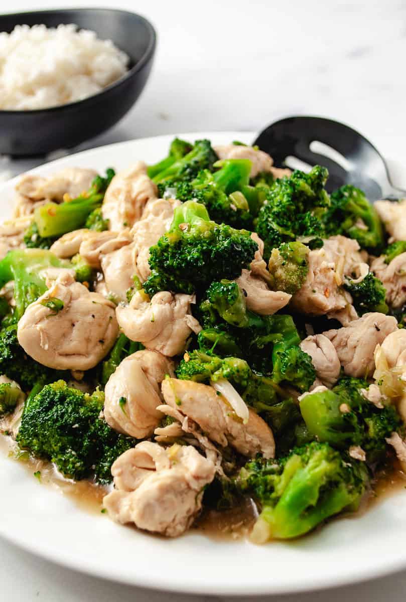 Easy Chinese Chicken Broccoli Better Than Takeout close up of chicken and broccoli on a white serving plate with a black slotted spoon, black bowl of white rice in the background.