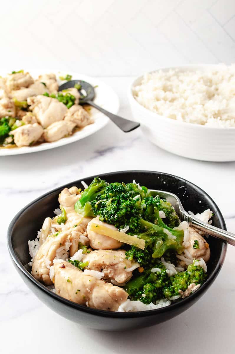 Easy Chinese Chicken Broccoli Better Than Takeout black round dish with the chicken and broccoli over white rice, a bowl of white rice in the top right and a white platter with the recipe top left with a black serving spoon.