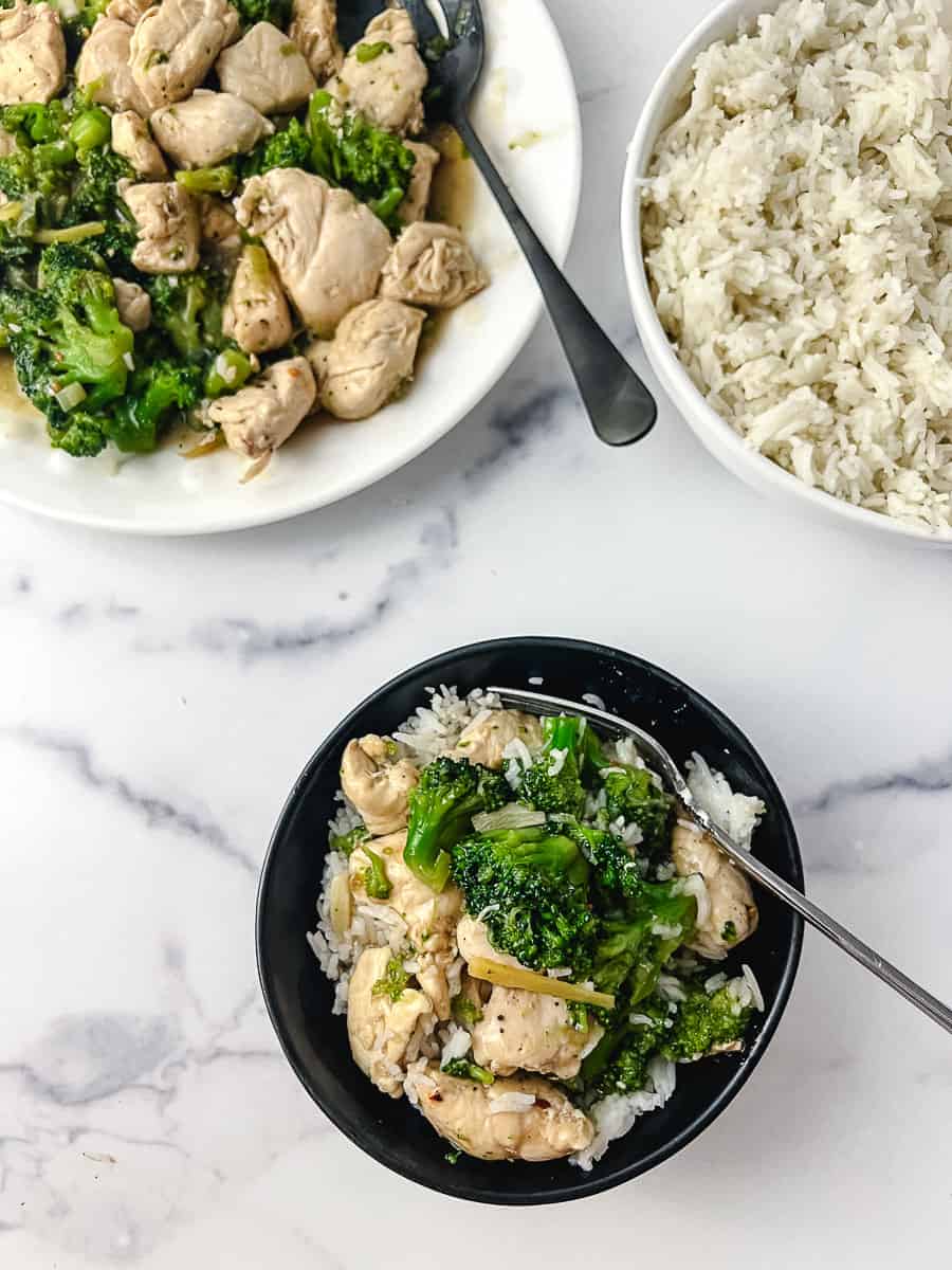 top down image of Easy Chinese Chicken Broccoli Better Than Takeout black round dish with the chicken and broccoli over white rice, a bowl of white rice in the top right and a white platter with the recipe top left with a black serving spoon.