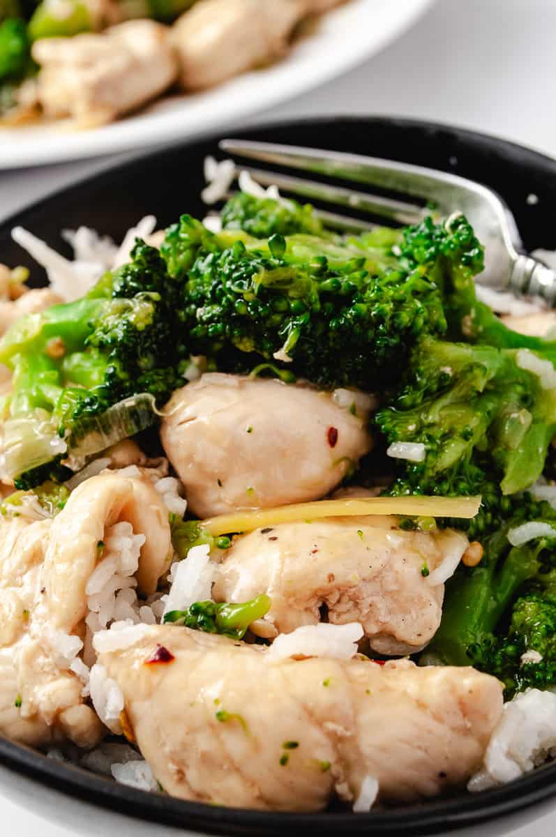 close up of the chicken and broccoli over white rice with a sliver of ginger in a black bowl and a fork in the blow.