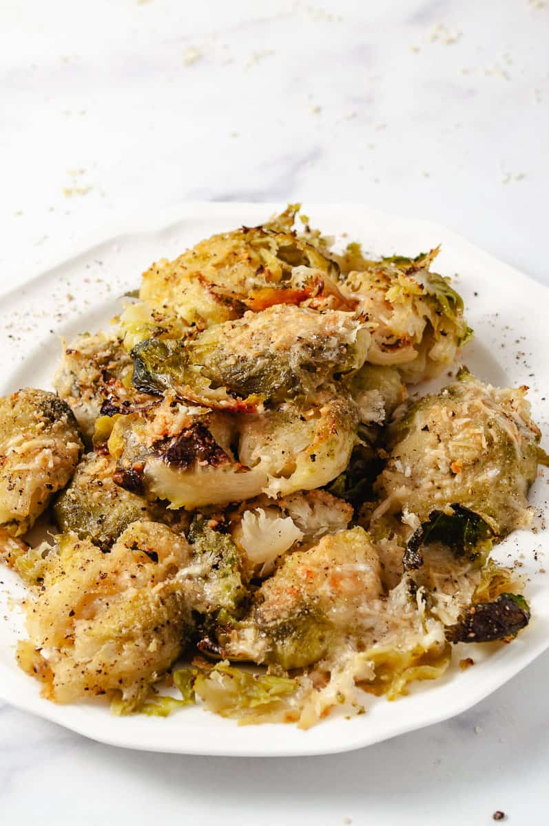 Easy Crispy Parmesan Smashed Brussels Sprouts 12 crispy brussels sprouts heaped on a white plate on a white counter, salt and pepper on the sprouts and plate.