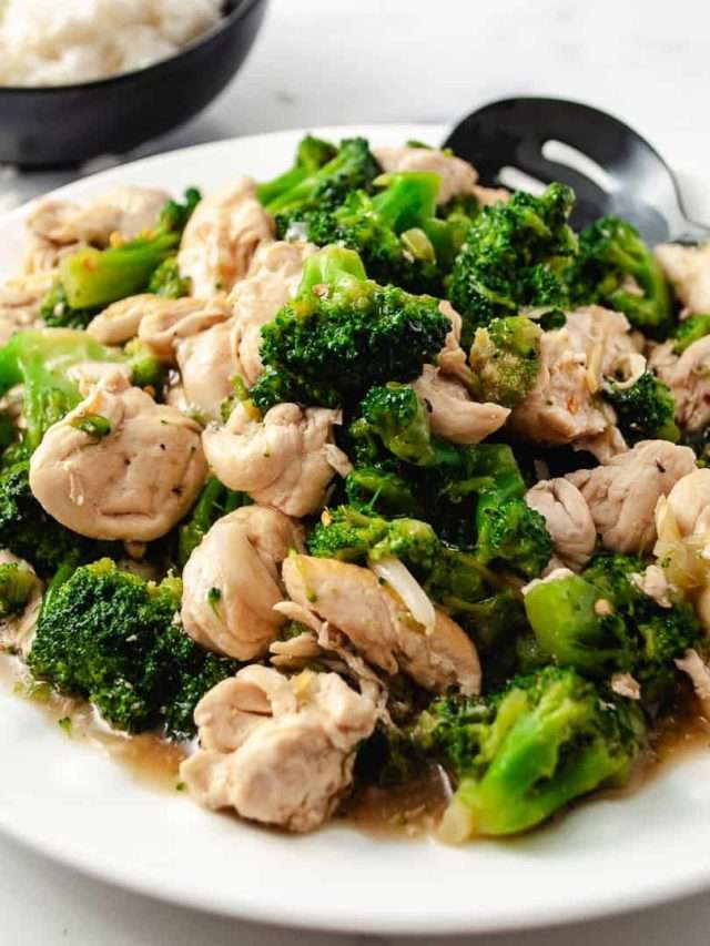 Easy Chinese Chicken Broccoli Better Than Takeout close up of chicken and broccoli on a white serving plate with a black slotted spoon, black bowl of white rice in the background.