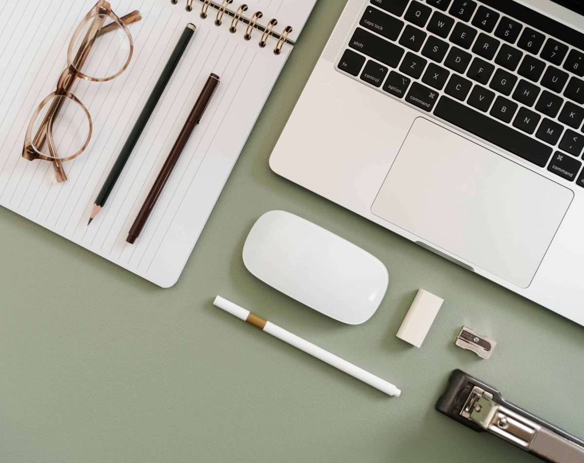 a green desk top that has on it a notebook, glasses a pen and pencil, a laptop, mouse pencil sharpener and a stapler.