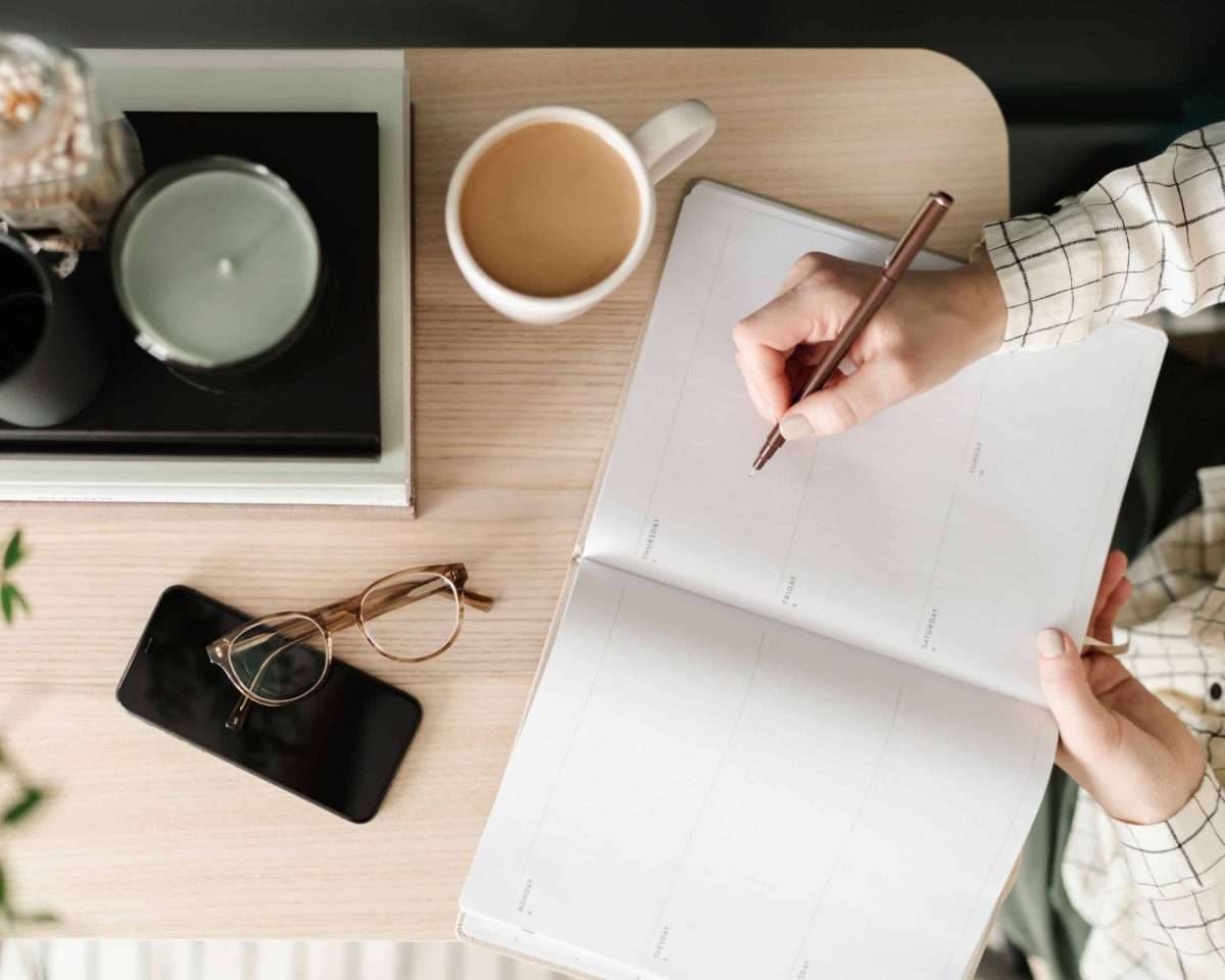 woman in a white shirt with black plaid writing in her calender top down on a blonde table with coffee cup a pair of glasses and a candle and 2 books