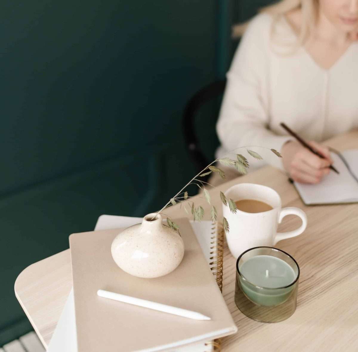 woman in a white sweater writing in a notebook her expenses, on the table a coffee cup a green candle 2 more notebooks, a pencil and a vase with a green spring.