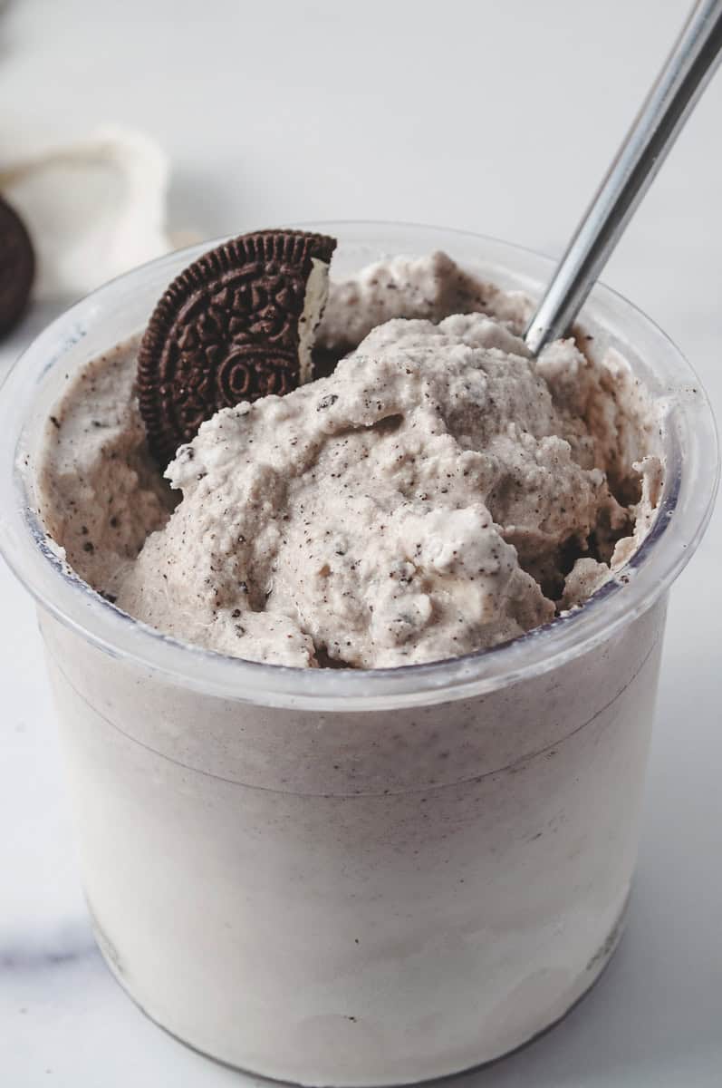 ninja creami cookies and cream with oreos ice cream mixed in and a cookie on top and a spoon, with an oreo on the counter.