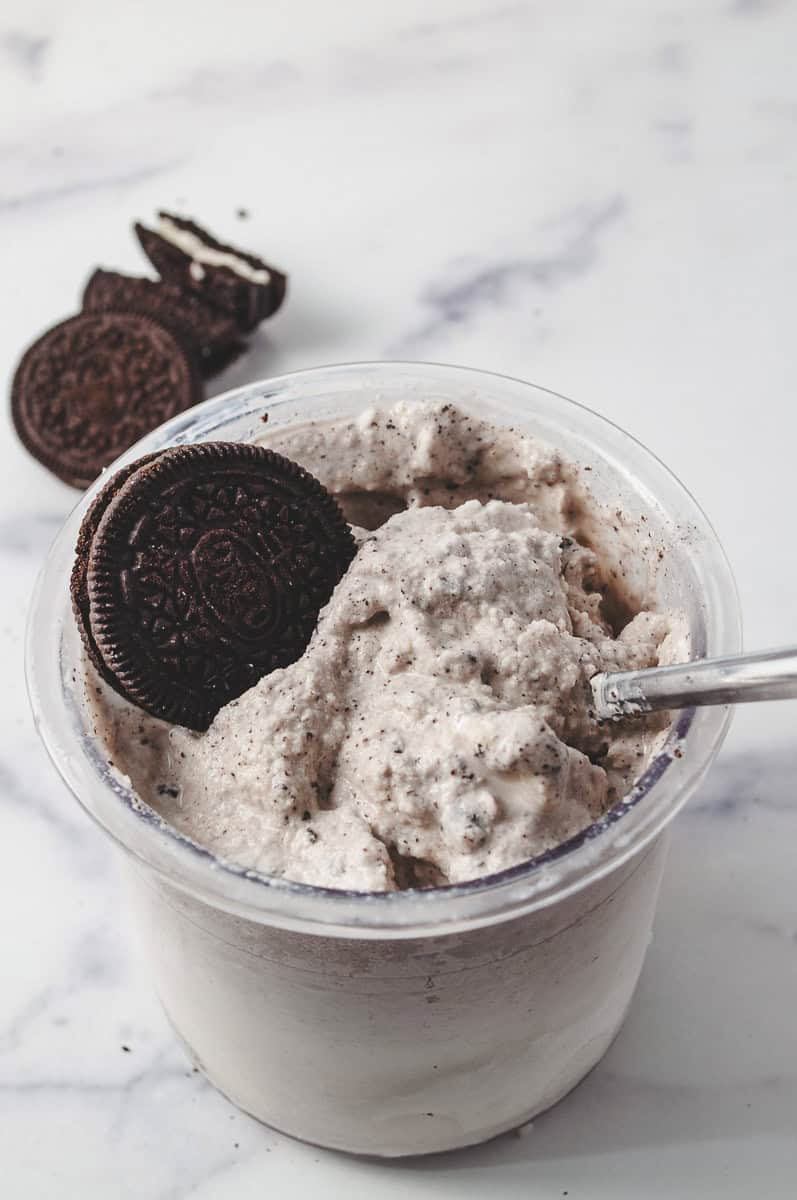 Ninja creami pint jar with the cookies and cream ice cream spun in the ninja cream machine and one oreo in the pint jar and a spoon in the jar and 3 cookies on the counter.