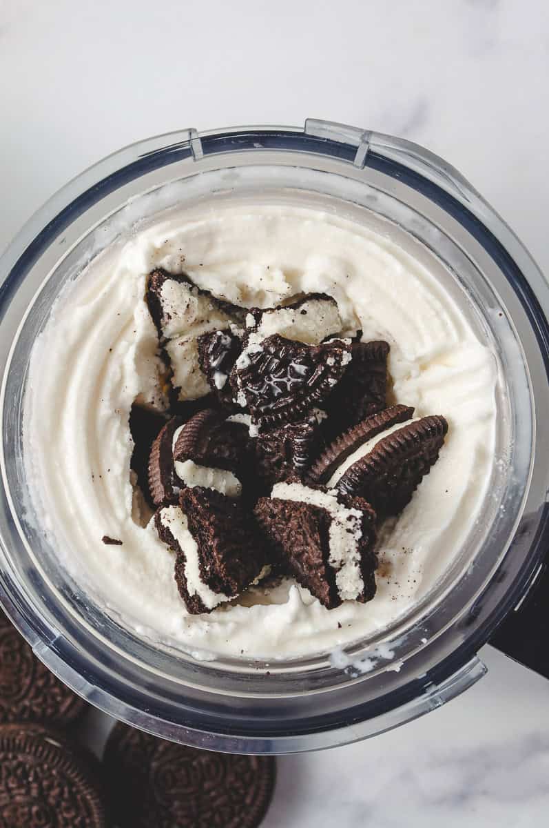top view of ninja creami cookies and cream pint jar with vanilla base ice cream and oreo cookie pieces on top and oreo cookies on the table.