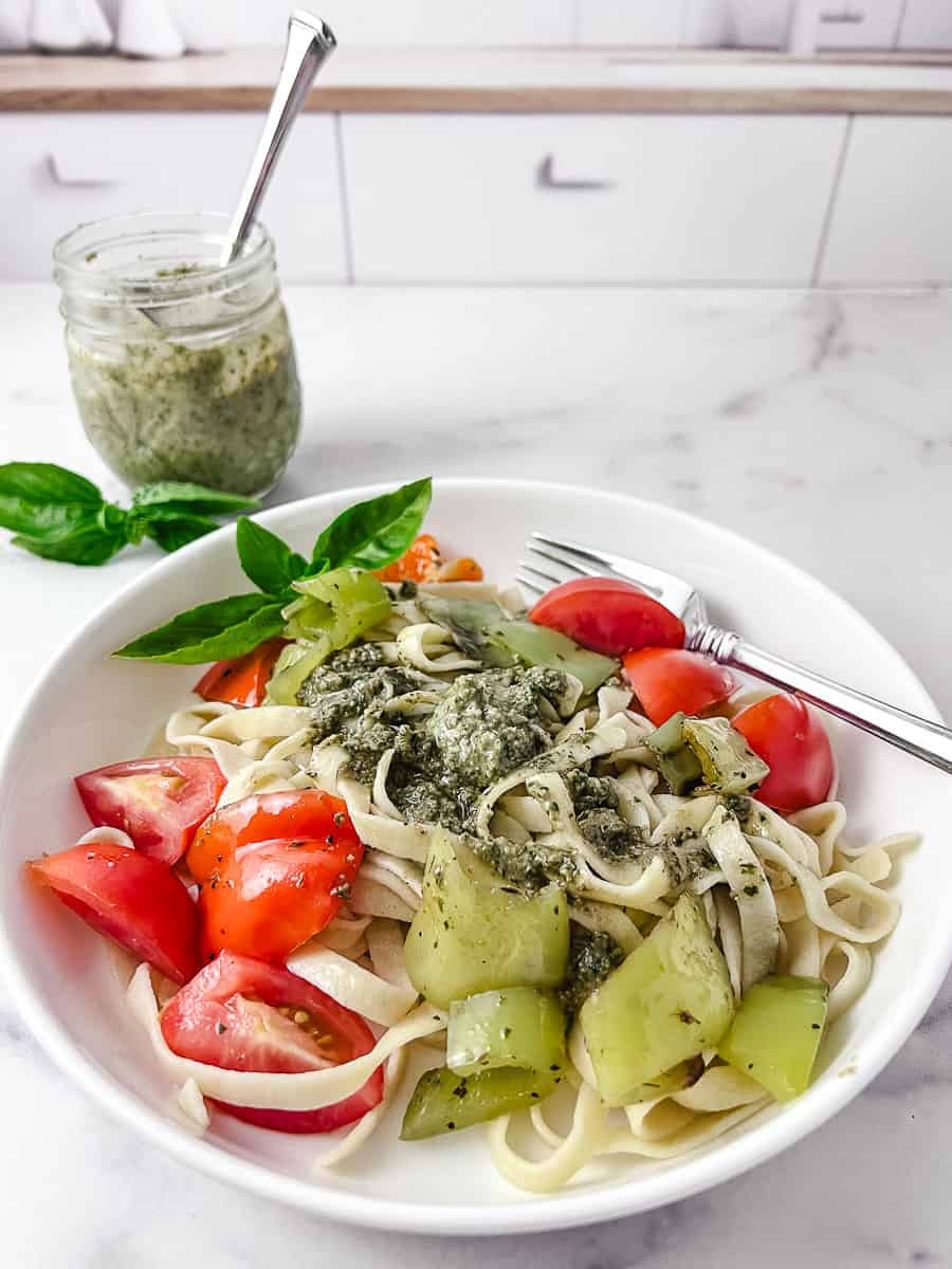 homemade fresh pesto with walnuts on homemade pasta with peppers and tomatoes on a white plate with a little mason jar of pesto in the top left.