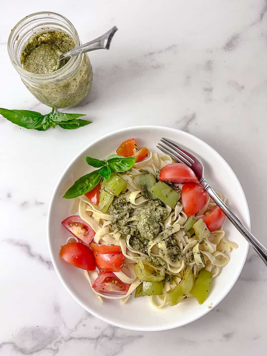 Homemade Fresh Italian Basil Pesto with Walnuts to down view of the plate with the pesto on homemade pasta with sauteed tomatoes and peppers and the jar of pesto at the top left with basil on the plate and counter. 
