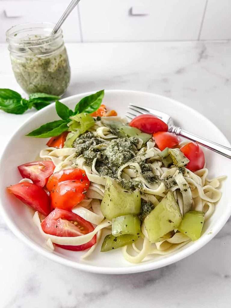 homemade fresh pesto with walnuts on homemade pasta with peppers and tomatoes on a white plate with a little mason jar of pesto in the top left.