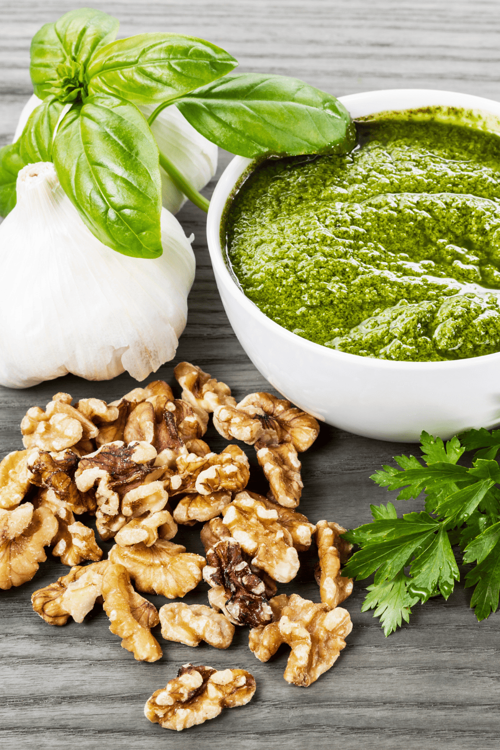 Homemade Fresh Italian Basil Pesto with Walnuts on a wood counter a white bowl of pesto 2 garlic bulbs, basil leaves and walnuts. 