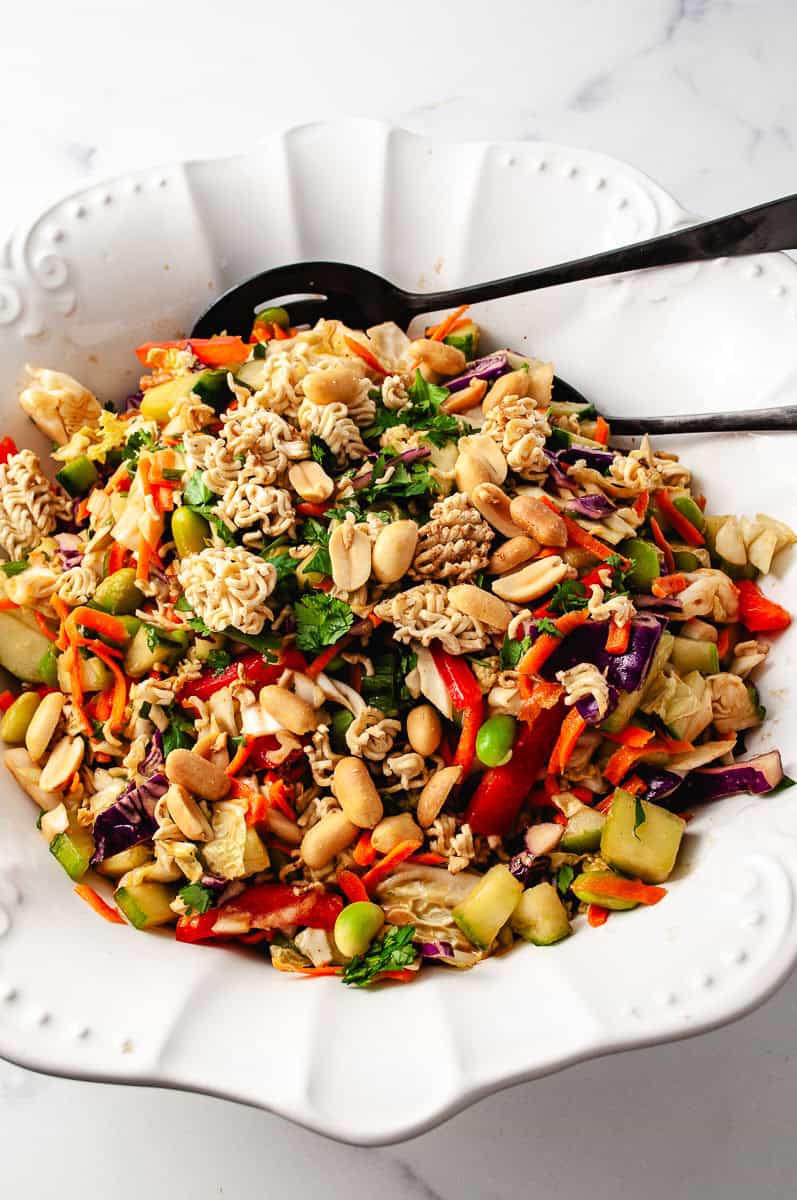 thai crunch salad close up of all the veggies with peanuts and ramen noodles on top in a white bowl with a black serving spoons.