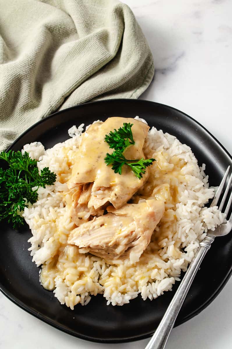 Creamy CrockPot Dry Ranch Dressing Chicken 5-Ingredients on a bed of rice on a black late with parsley spring on top of the chicken and the side of the dish with a fork and a green napkin top left.