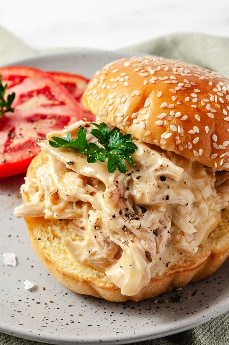 Creamy CrockPot Dry Ranch Dressing Chicken 5-Ingredients the chicken shredded on a bun with sesame seeds on the top bun and a sprig of parsley on the chicken and 2 sliced tomatoes all on a gray plate.