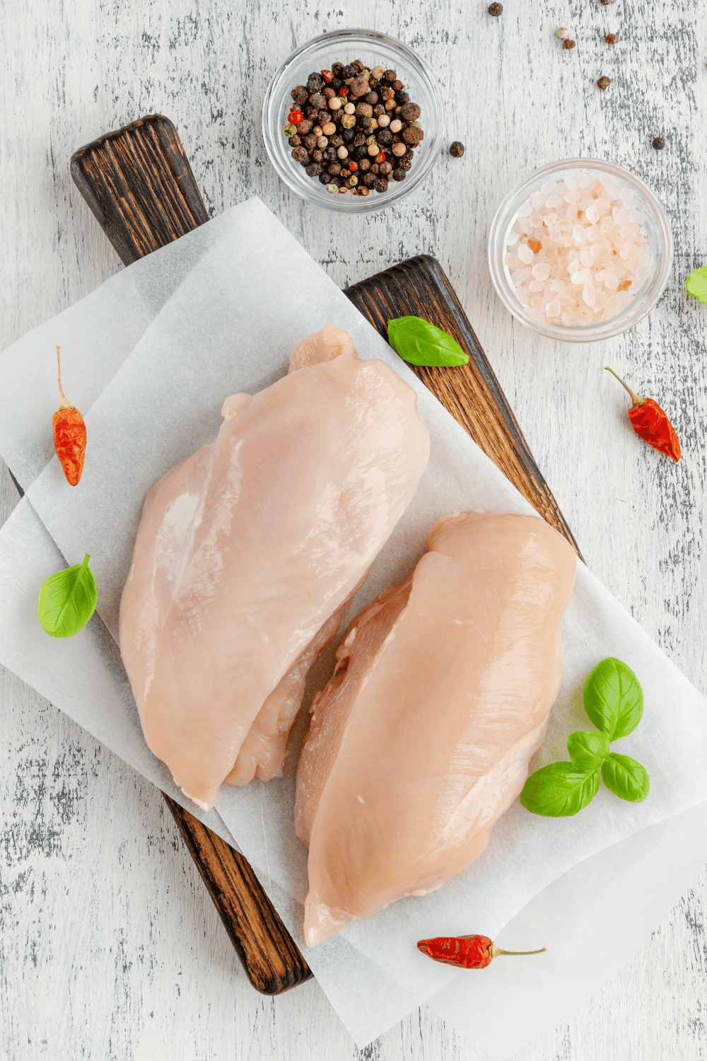 2 chicken breast raw on a cutting board with salt and pepper in small clear bowls.