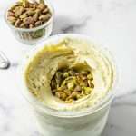 Ninja Creami Pistachio Ice Cream No Artificial Dyes mixed ice cream in the pint container with crushed pistachio in the center and a small clear bowl of whole shelled pistachios in the background on a white counter