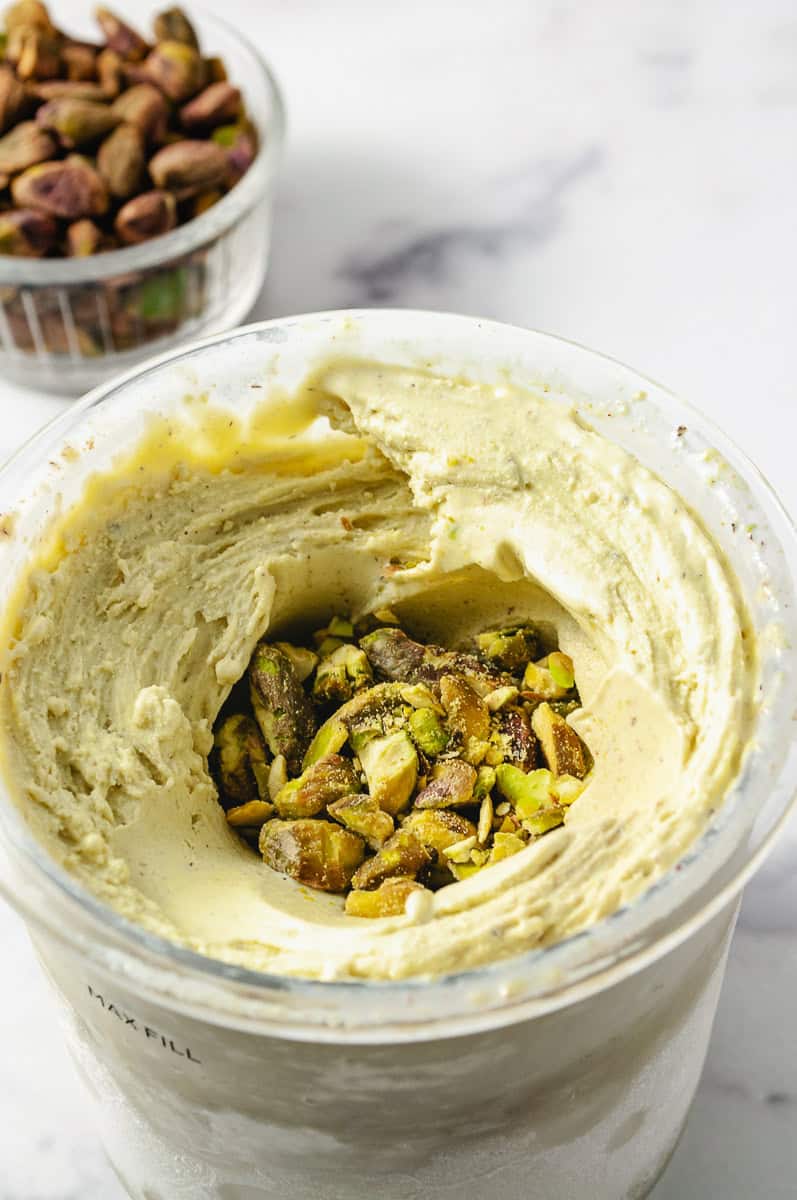 Ninja Creami Pistachio Ice Cream No Artificial Dyes mixed ice cream in the pint container with crushed pistachio in the center and a small clear bowl of whole shelled pistachios in the background on a white counter