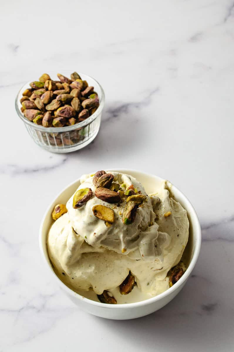 Ninja Creami Pistachio Ice Cream No Artificial Dyes mixed ice cream in the white bowl with crushed pistachio in the center and a small clear bowl of whole shelled pistachios in the background on a white counter