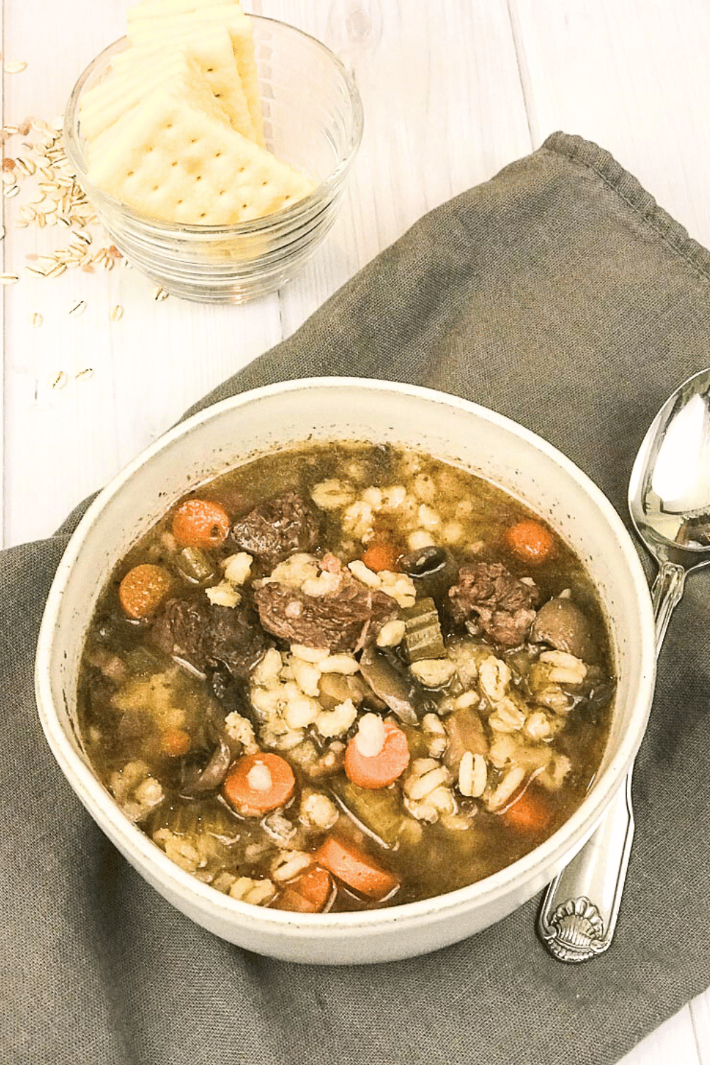 beef barley soup in a gray bowl on a gray napkin with a spoon and crackers in the top of the image.