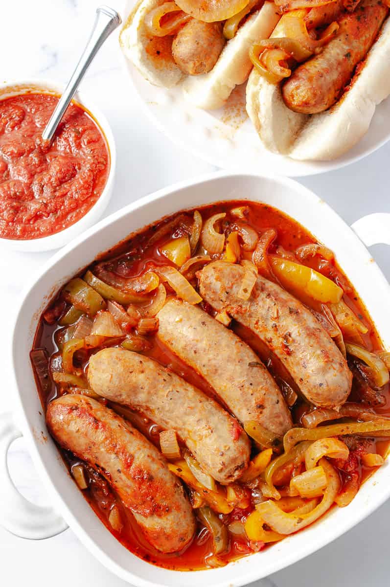 Easy CrockPot Italian Sausages Peppers Onions a white square serving dish with 4 sausages with the onion, pepper and tomato sauce a small white round dish with tomato sauce and a spoon and at the top right 2 sausages in two buns loaded with peppers and onions.