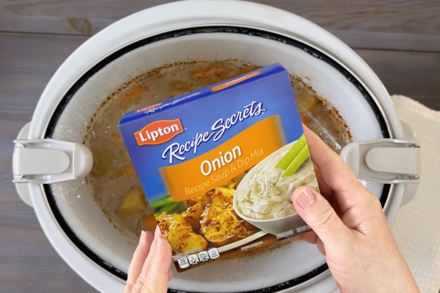 lipton soup mix box in womens hand holding over the white Crockpot.