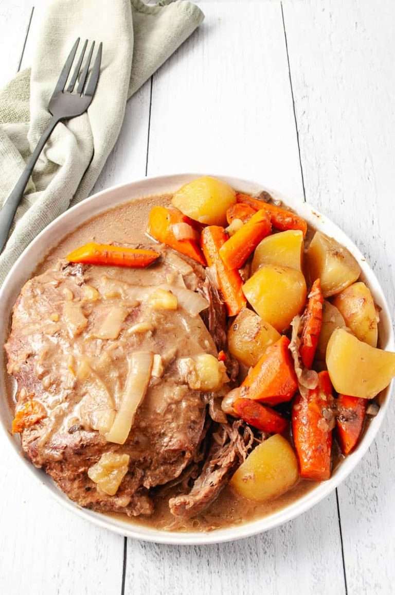 Easy Slow Cooker Pot Roast Lipton Onion Soup Mix on a round white plate with onions, carrots and potatoes with a green kitchen towel and a serving fork on a white wood table.