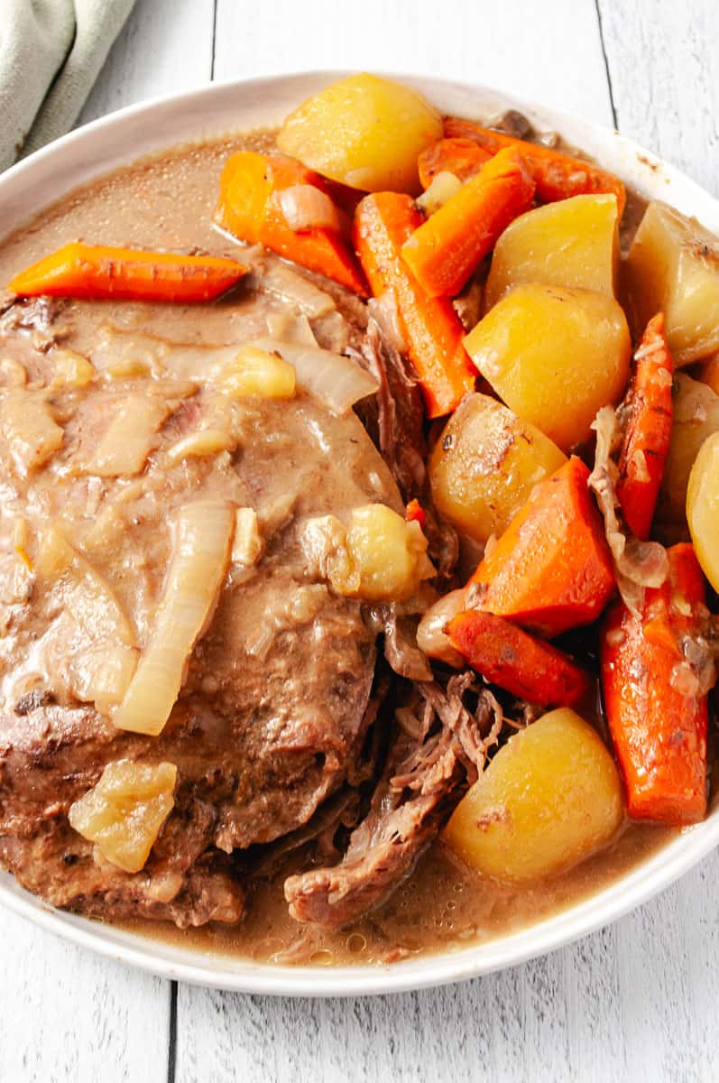 Easy Slow Cooker Pot Roast Lipton Onion Soup Mix on a round white plate with onions, carrots and potatoes on a white wood table close up of the roast.