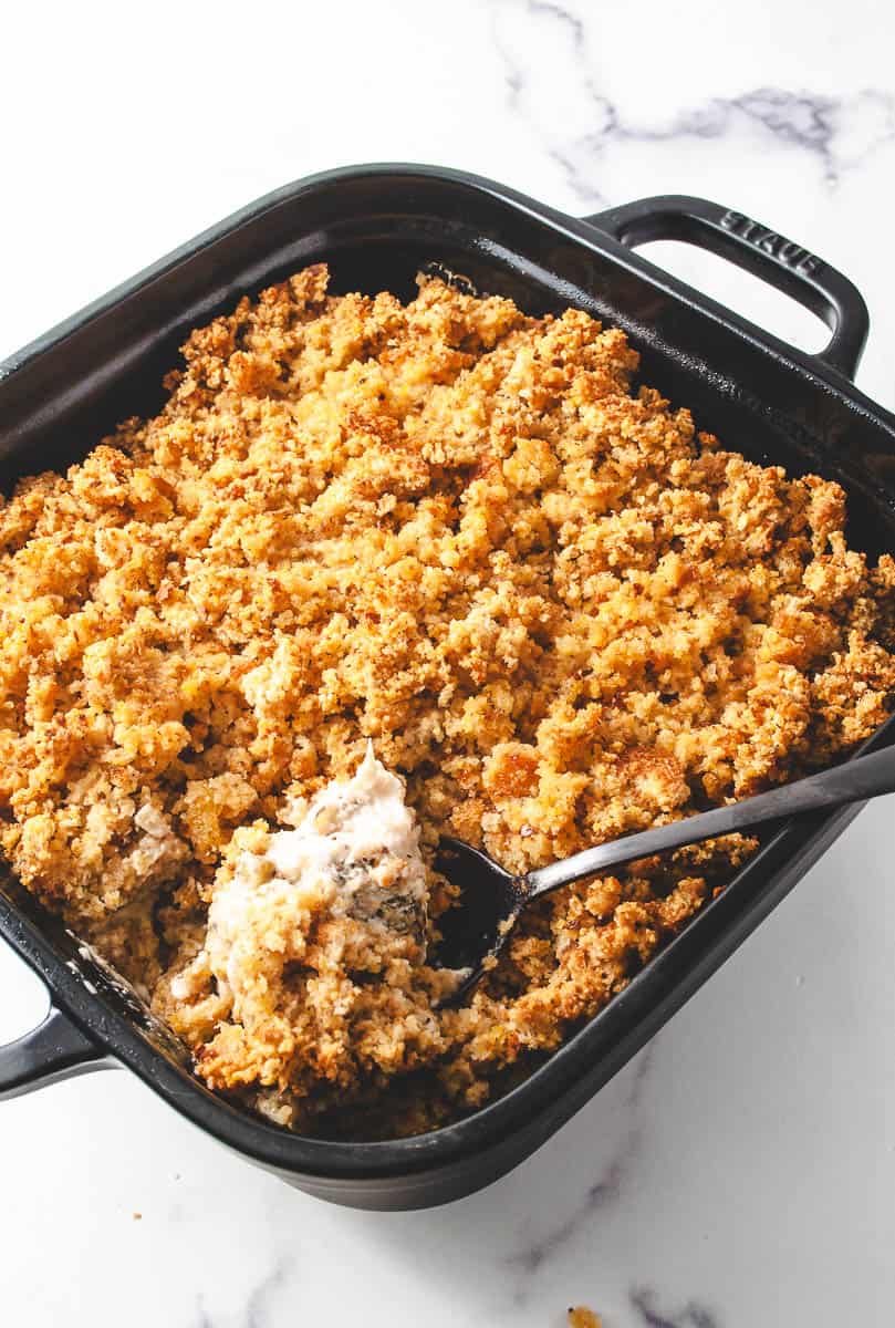 one dish chicken stuffing casserole in a black square cast iron dish on a white counter with a serving spoon lifting a chicken from the casserole.