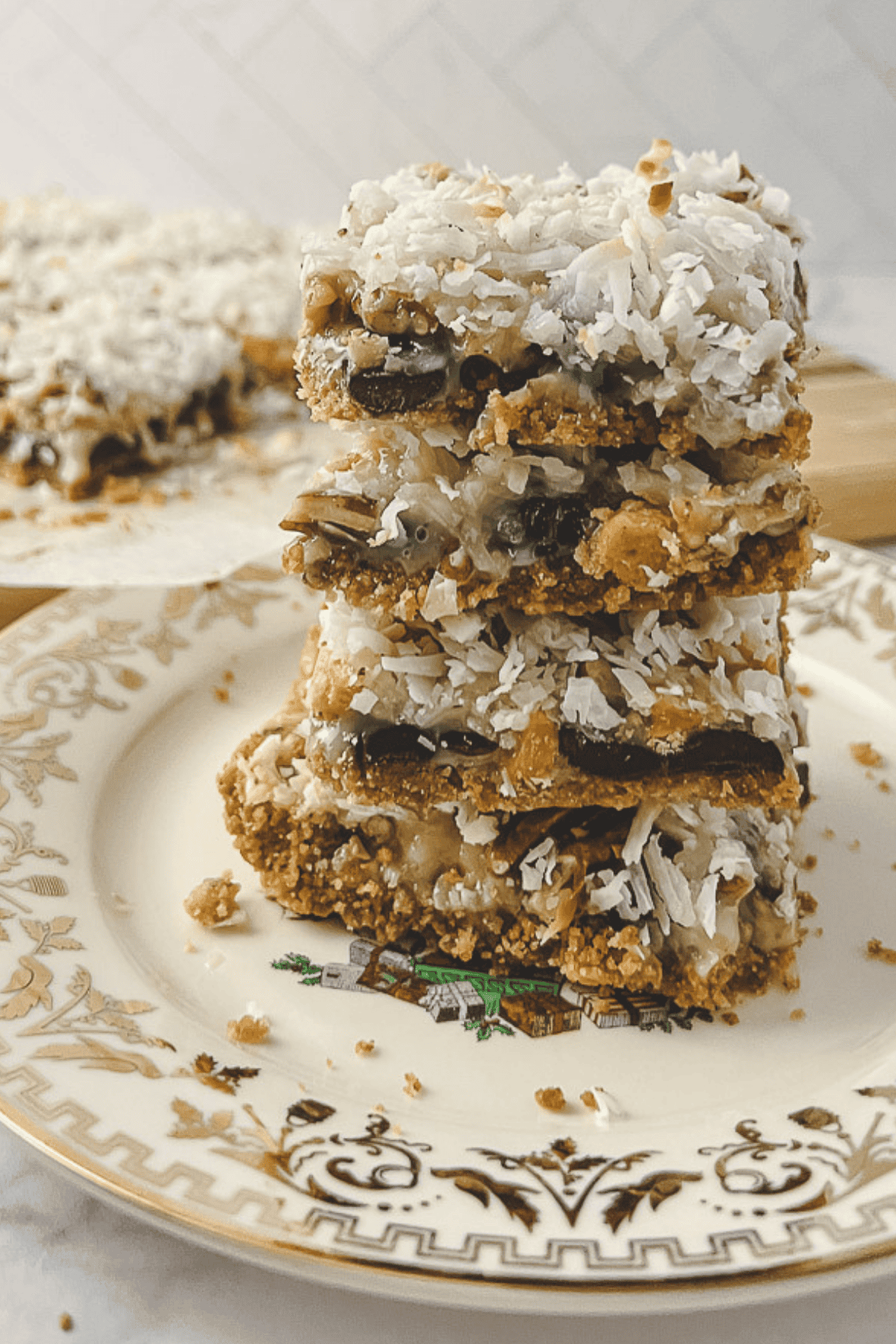 Stack of four magic cookie bars showing the graham cracker crust, chocolate, pecan and coconut layers on a gold flowered Christmas plate.