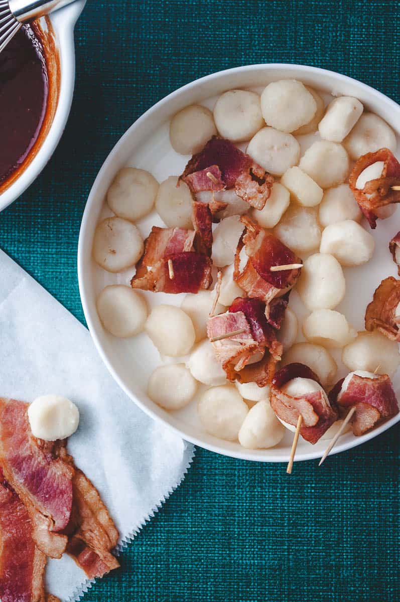Bacon Wrapped Water Chestnuts with Pre-Cooked Bacon (Rumaki) top down view of the water chestnuts in a white plate being wrapped with bacon.