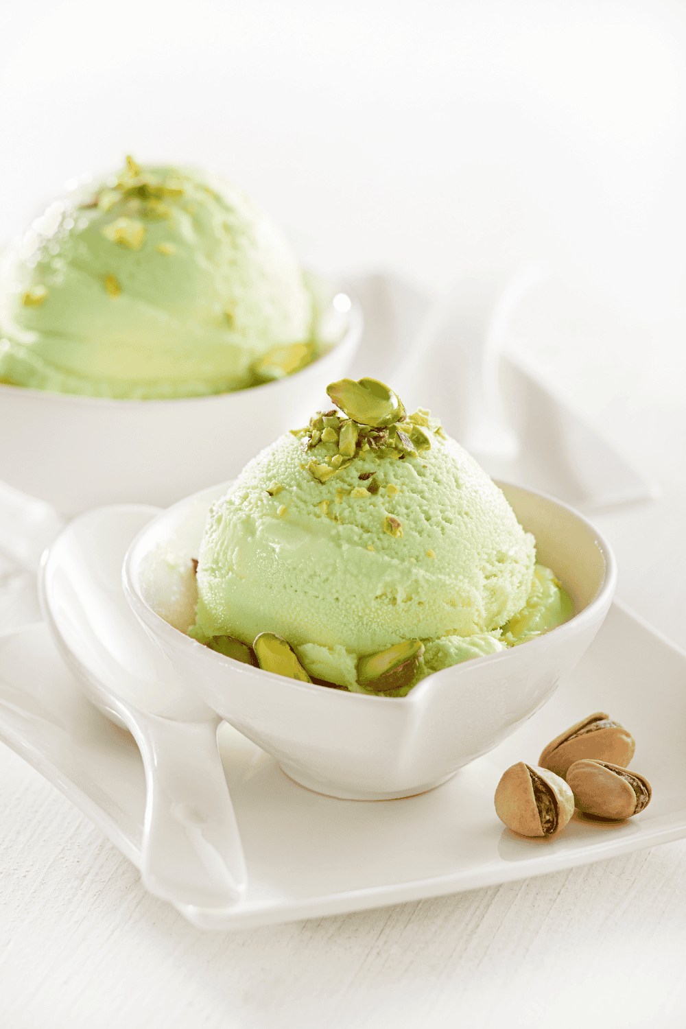 Pistachio Ice Cream Ninja Creami 2 bowls of ice cream white on a white tray with a few pistachio nuts in their shells and a few crushed nuts crushed ont op of the ice cream.