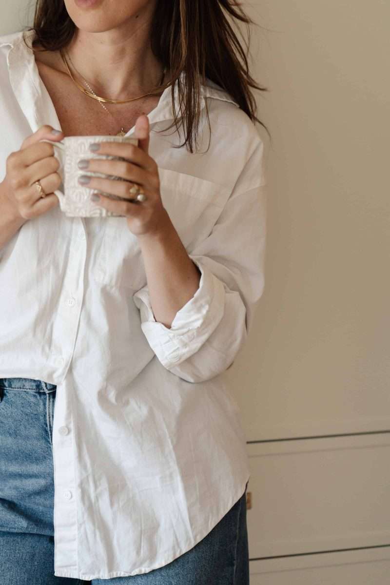 Using the Envelope System: A Beginner’s Budgeting Guide a woman and a white shirt holding a white coffee cup in blue jeans against a tan wall.
